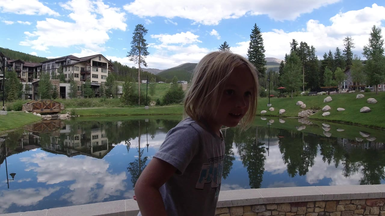 The girl remains near the bridge edge in Winter Park, Colorado, with the lake reflecting clouds and nearby lodge buildings across the water.