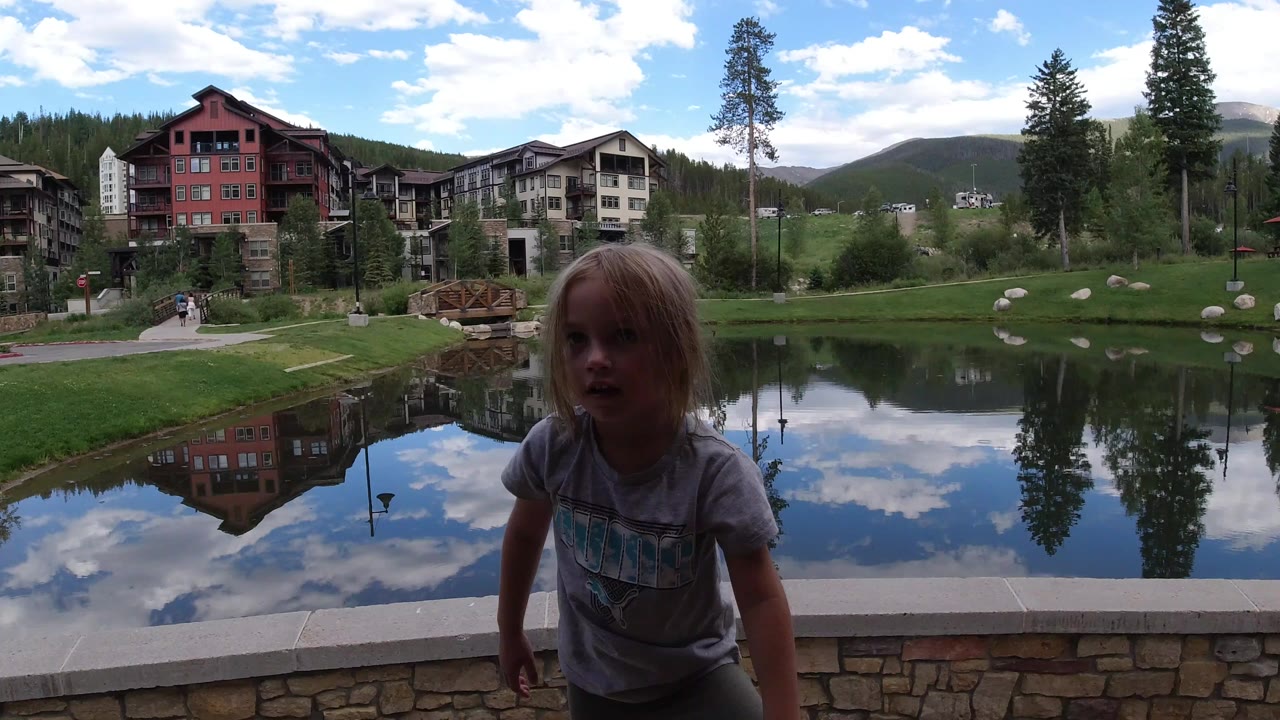 A young girl faces the camera in Winter Park, Colorado, standing by a stone lakeside edge with reflected clouds, trees, and houses behind her.