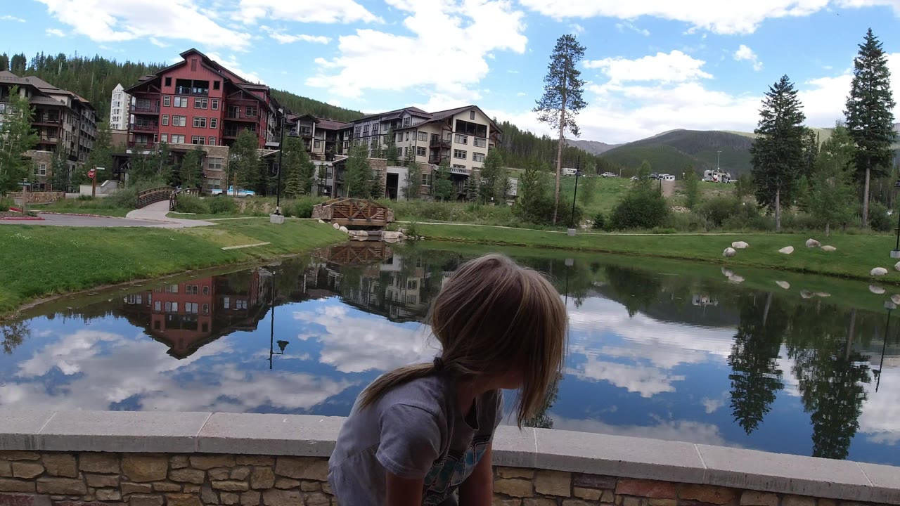 The girl remains near the stone edge as a red building, adjacent lodging, trees, blue sky, and white clouds reflect clearly in the pond at Winter Park, Colorado.