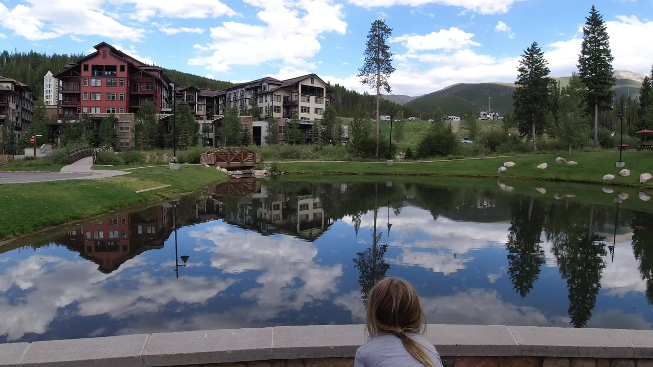 Wide Winter Park, Colorado pond view with lodge-style buildings, evergreen trees, cloudy blue sky, and the back of a blonde child low in the foreground.