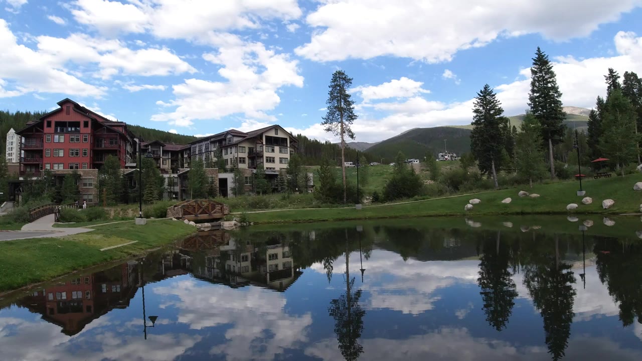 Pond reflecting lodge buildings, trees, clouds, and hills.