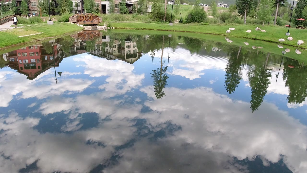 Winter Park, Colorado lake reflection showing blue sky and clouds across the water, with shoreline houses, tall trees, lawn, rocks, a bridge, and a distant person near the upper left.