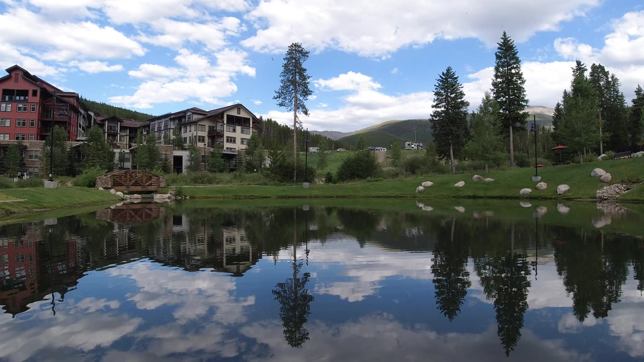Pond reflecting buildings, trees, clouds, and mountains.