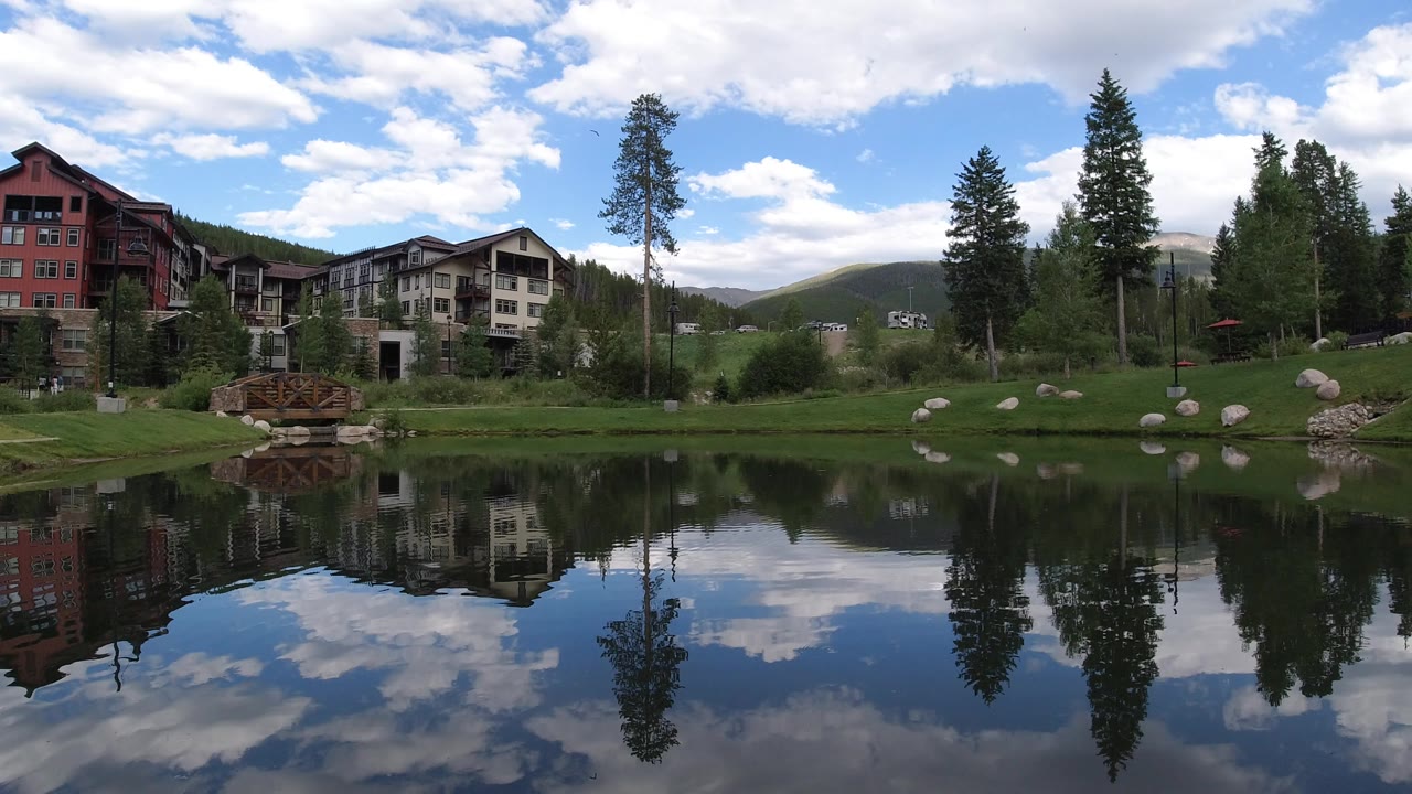 Winter Park, Colorado pond scene with lodge-style buildings at left and center, tall trees across the lawn, cloud reflections, and no visible people.