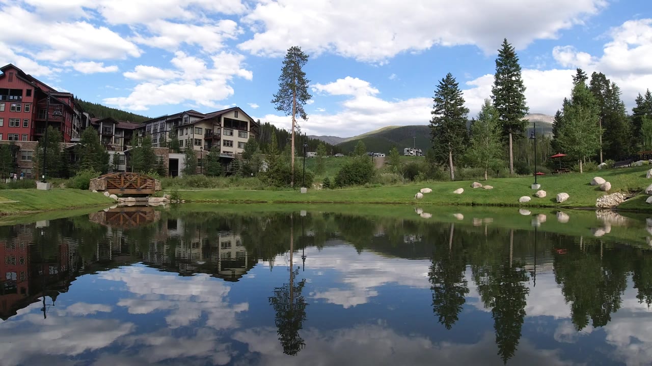 Wide Winter Park, Colorado pond view at the start of the clip, with lodge-style buildings, tall evergreens, a grassy bank, boulders, a small wooden footbridge, cloud reflections, and no visible people.
