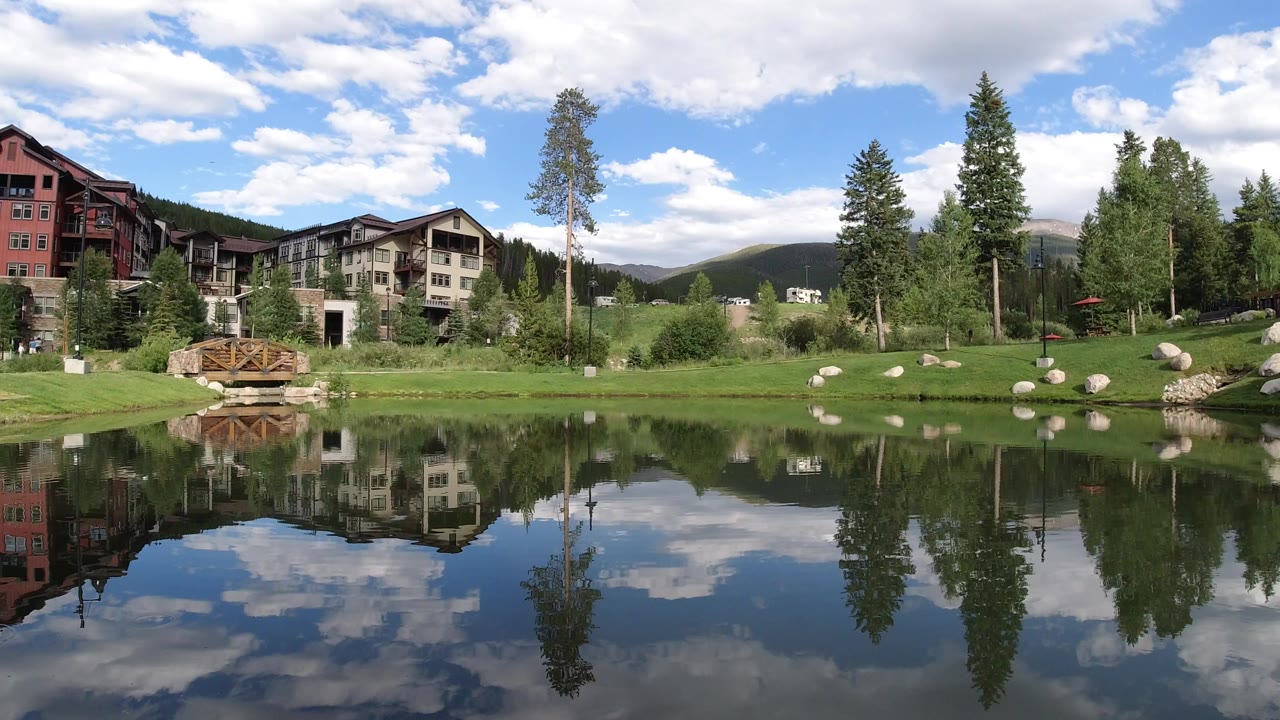 Wide Winter Park, Colorado lake view at the start of the clip, with multi-story buildings, a wooden bridge, trees, mountains, and cloud reflections in the water, no visible people.