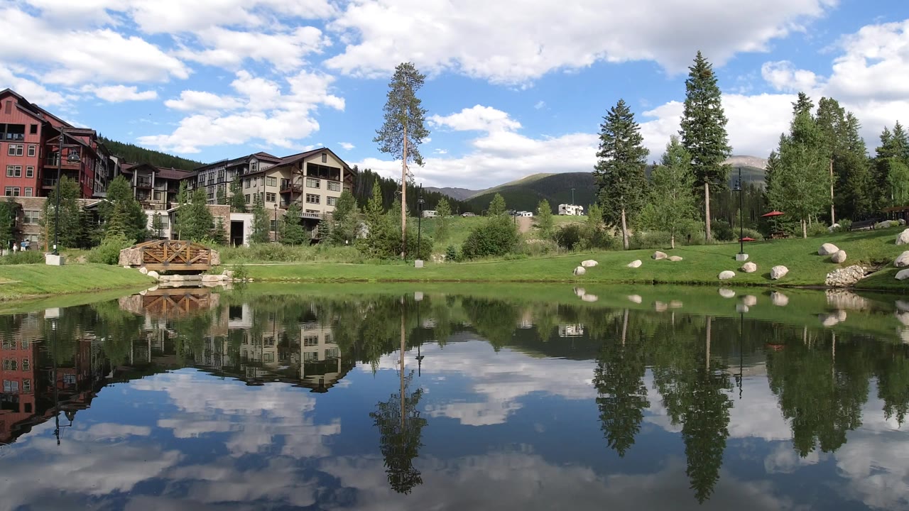Winter Park, Colorado lakefront frame with the wooden bridge near the left center, lodge-like buildings behind it, tall trees, distant mountains, and rippled cloud reflections, no visible people.