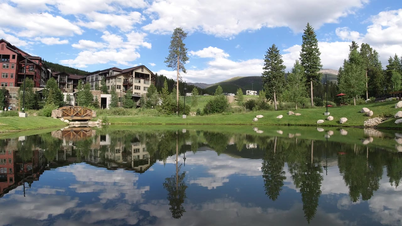 Lake, landscaped shore, tall trees, buildings, and cloudy sky reflections.