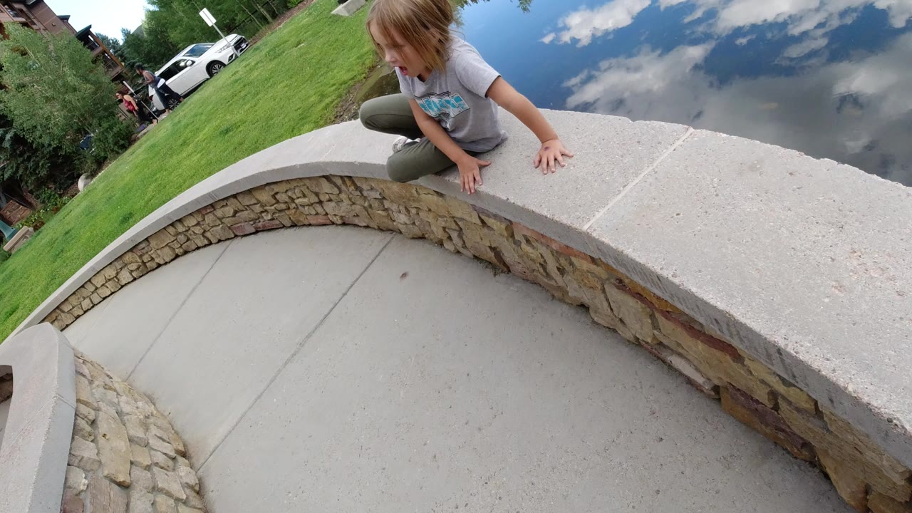 Young girl crouches on a curved stone wall beside a reflective lake.