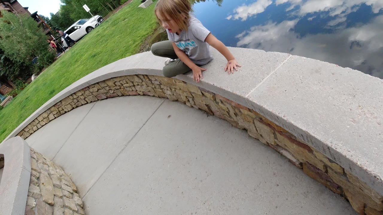 In Winter Park, Colorado, the girl remains near the top of the curved stone wall, looking down toward the walkway as the still lake reflects blue sky and clouds.