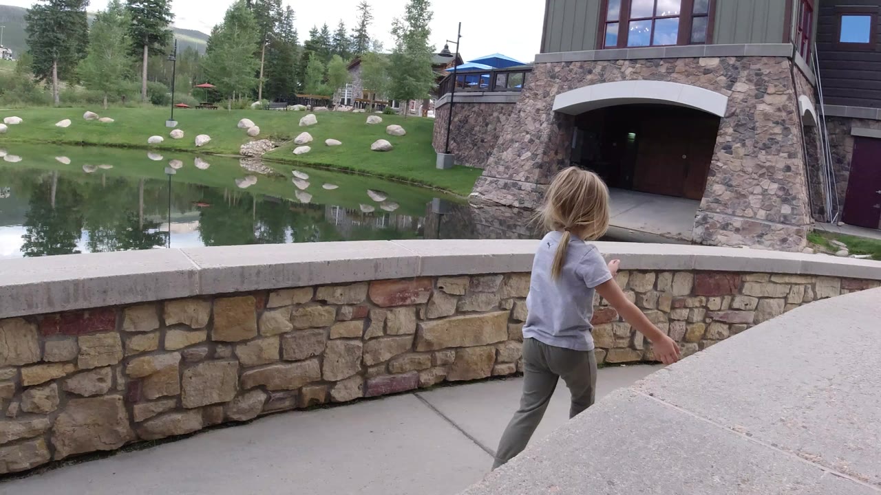 A young girl walks along a stone bridge wall beside a reflective pond in Winter Park, Colorado, with pine trees, boulders, lawn, and a building entrance in the background.
