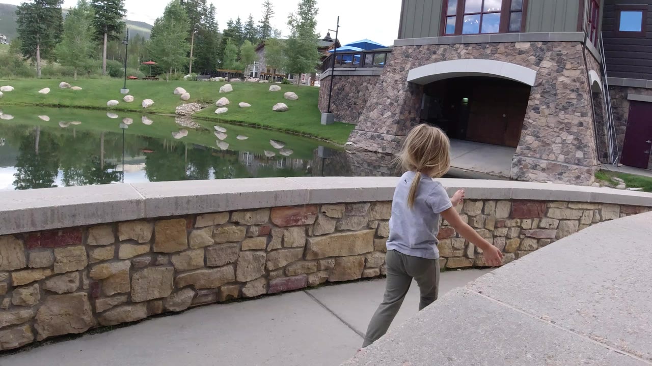 The young girl moves farther along the low stone wall in Winter Park, Colorado, with the pond, evergreen trees, rocks, blue umbrellas, and stone-faced building visible behind her.
