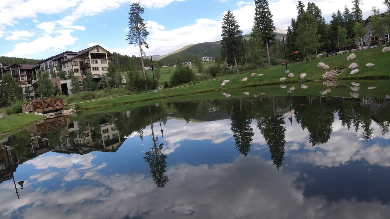 Winter Park, Colorado pond scene with apartment-style homes on the left, a narrow wooden bridge near the shore, scattered rocks on the grass, and trees reflected in dark water.