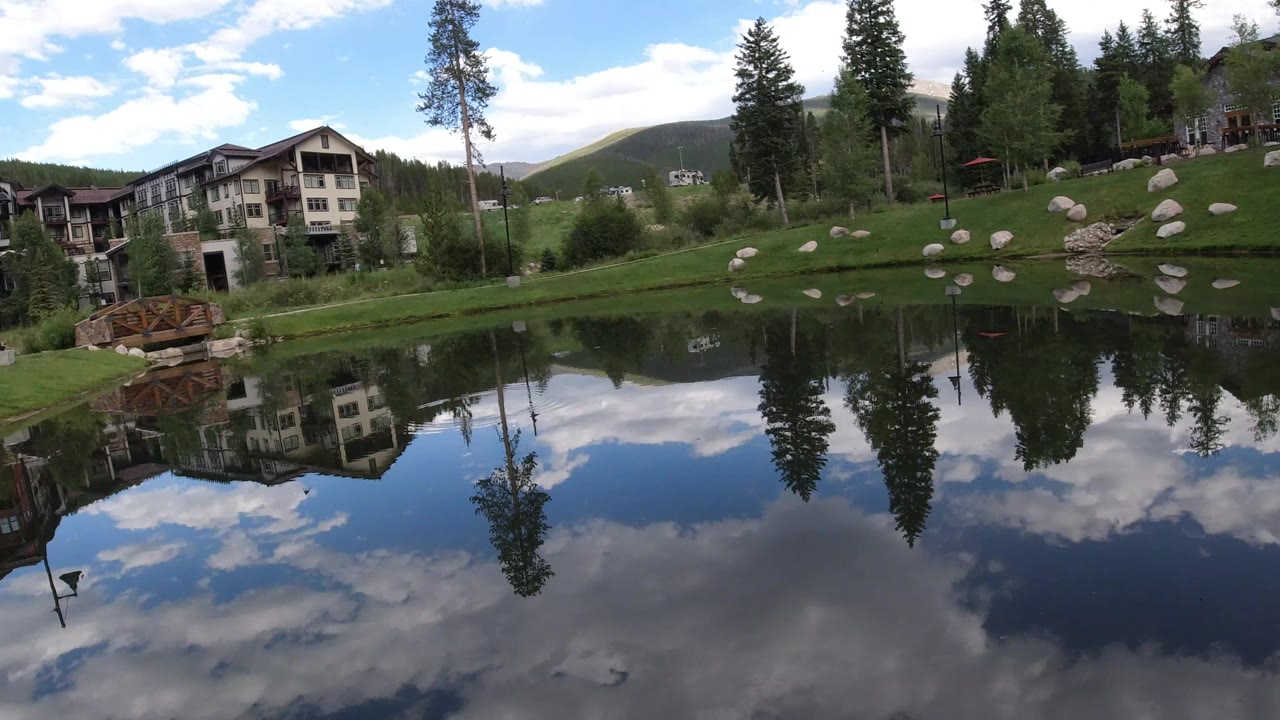 At 0:00 in Winter Park, Colorado, a wide pond reflects clouds, tall trees, apartment buildings, a wooden bridge, grass, and scattered boulders.