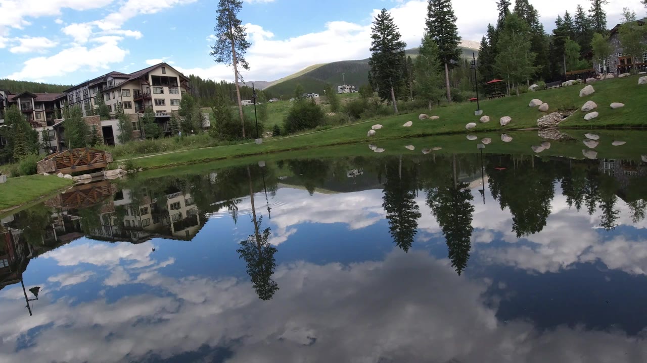 At 0:01 in Winter Park, Colorado, the pond surface mirrors a patch of blue sky while buildings, trees, grass, boulders, and a small bridge sit across the bank.
