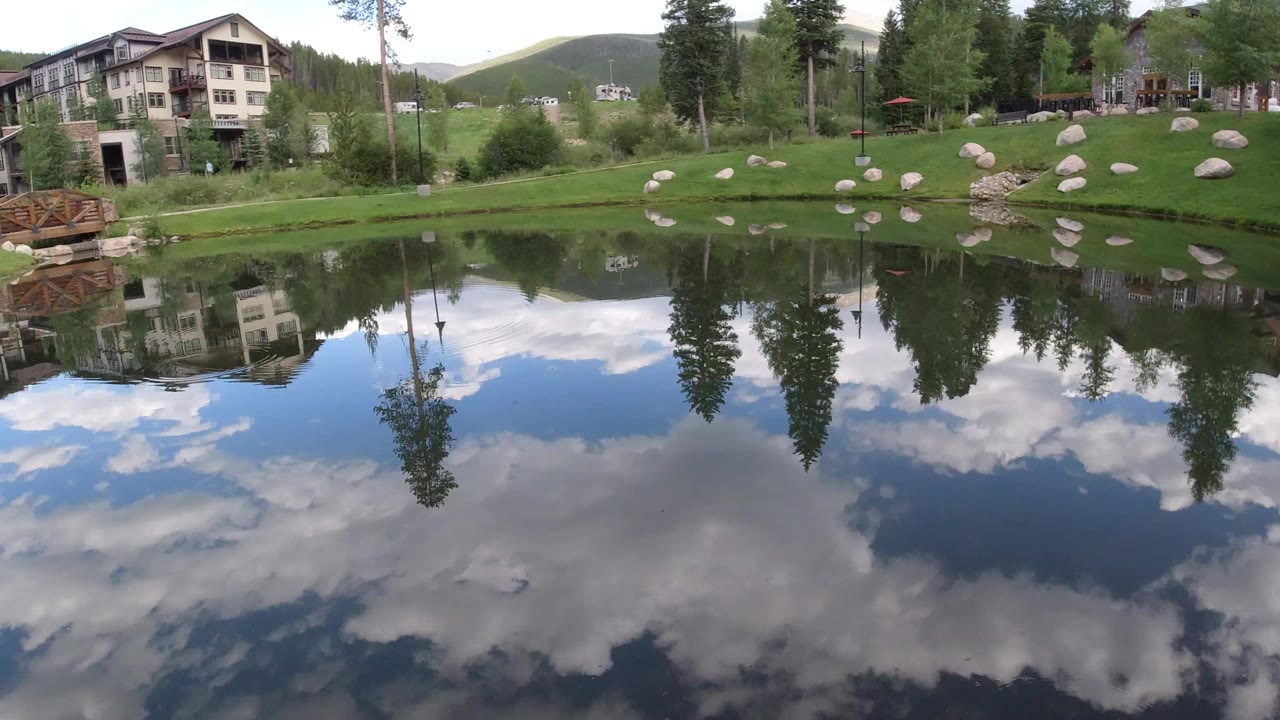 Pond reflecting lodge buildings, trees, clouds, and green lawn