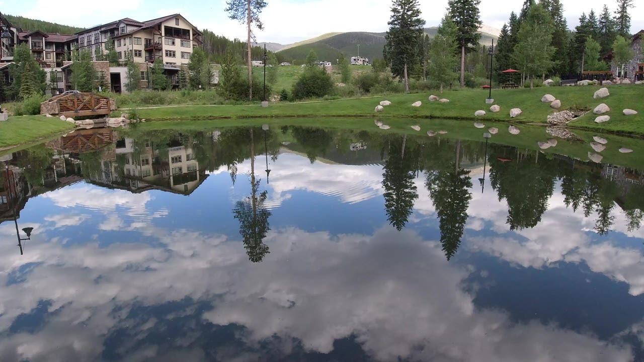 Winter Park, Colorado pond view with lodge-style buildings at left, evergreen trees, a wooden bridge, cloud reflections on the water, and no visible people.