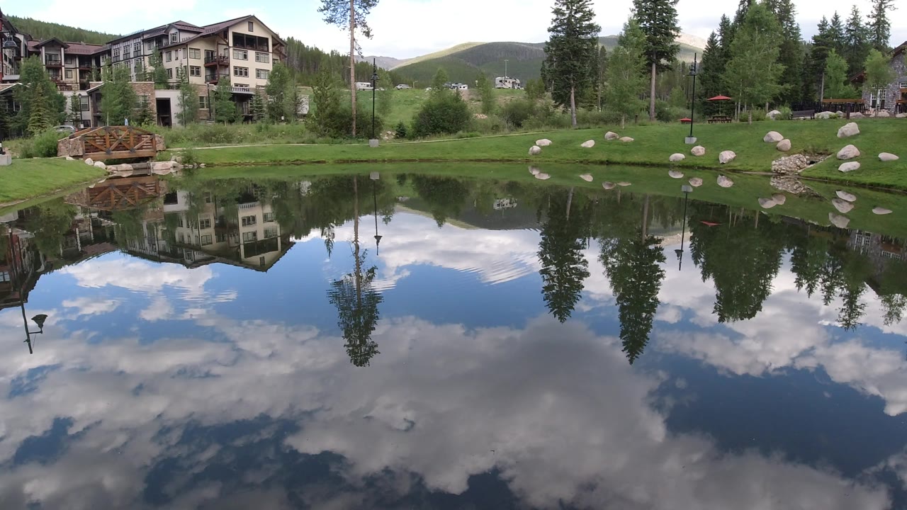 Pond reflection with tall trees, hillside, rocks, and lodge buildings.