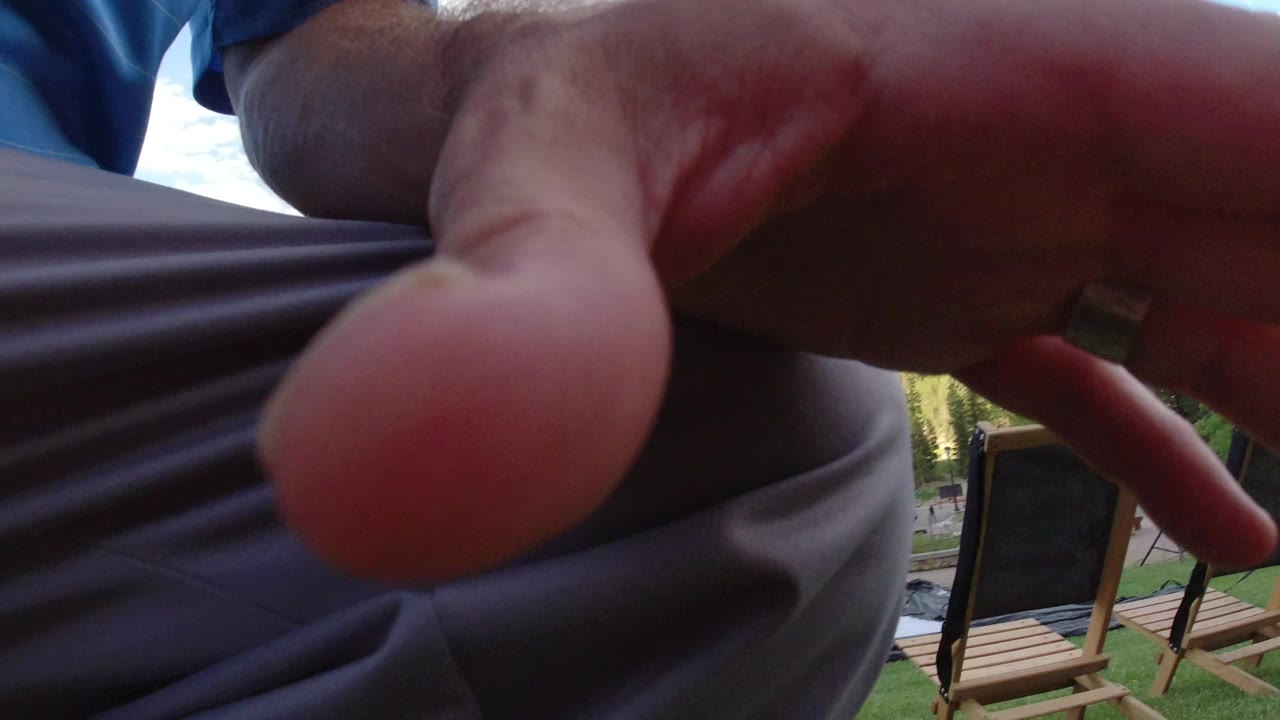 At 0:01 in Winter Park, Colorado, a finger fills the center foreground while wooden deck chairs, green turf, and bright outdoor background remain partly visible.