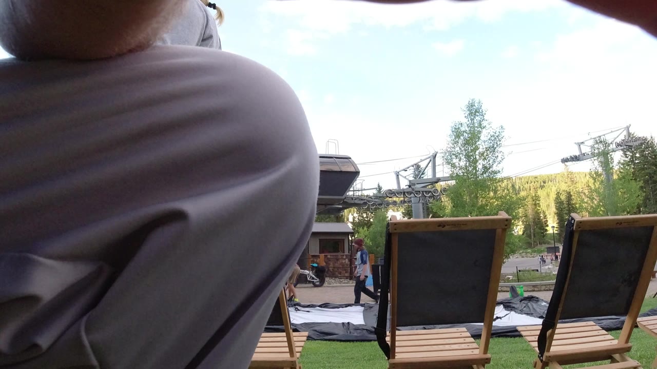 A close foreground leg in light pants dominates the left side while two wooden lawn chairs, a grassy area, trees, sky, pedestrians, and a dark roofline sit in the background in Winter Park, Colorado.