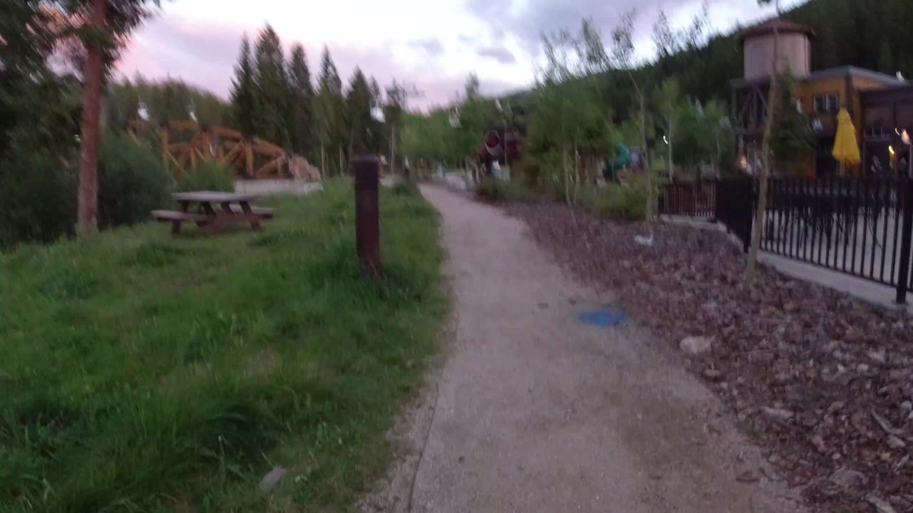 Start of a ground-level view along a gravel path in Winter Park, Colorado, with grass on the left, black fencing and lodge buildings on the right, and no visible people.