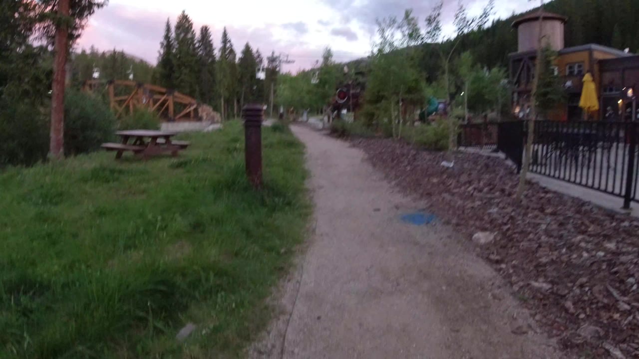 A similar Winter Park, Colorado path view shows a picnic table and dark bollard nearer the center, with black fencing and yellow umbrellas to the right and no visible people.