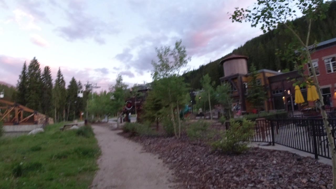 A Winter Park, Colorado walkway leads through landscaped greenery toward a wooden bridge and red building, with a small person visible near the path.