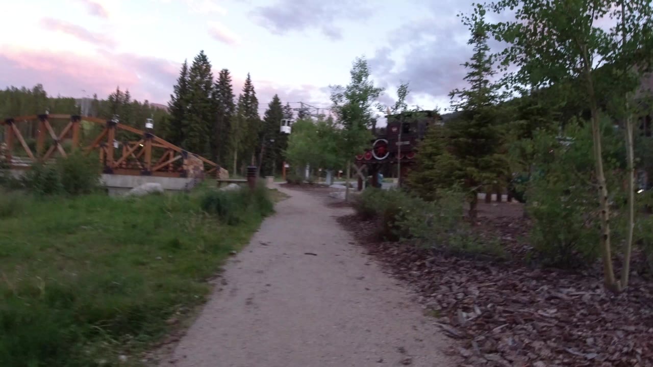 A Winter Park, Colorado gravel path curves past grass and shrubs toward a wooden bridge, with evergreen trees and pink evening clouds in the background and no visible people.