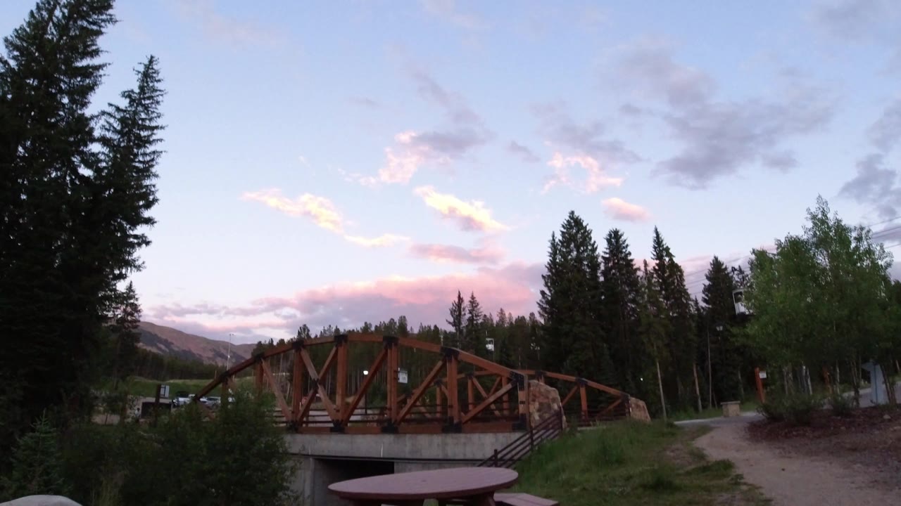 Opening frame of a rust-colored bridge in Winter Park, Colorado, with tall evergreens, grass, a gravel path, distant hills, pink clouds, and no visible people.