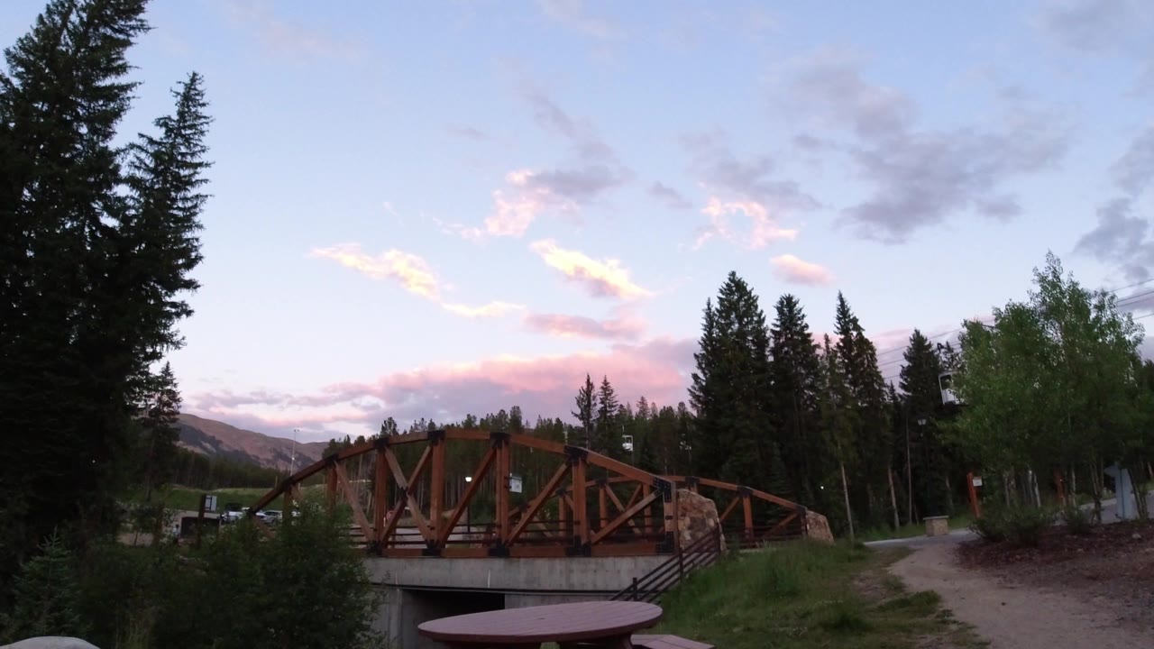 The bridge remains centered in Winter Park, Colorado, with dark conifers on both sides, a pale blue sky with pink clouds, grass, path, and no visible people.