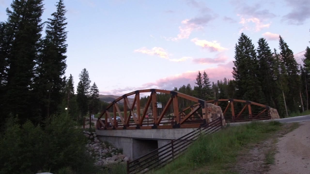Rust-colored wooden bridge in Winter Park, Colorado, crossing a concrete culvert beside a road, with dense evergreens and no visible people.