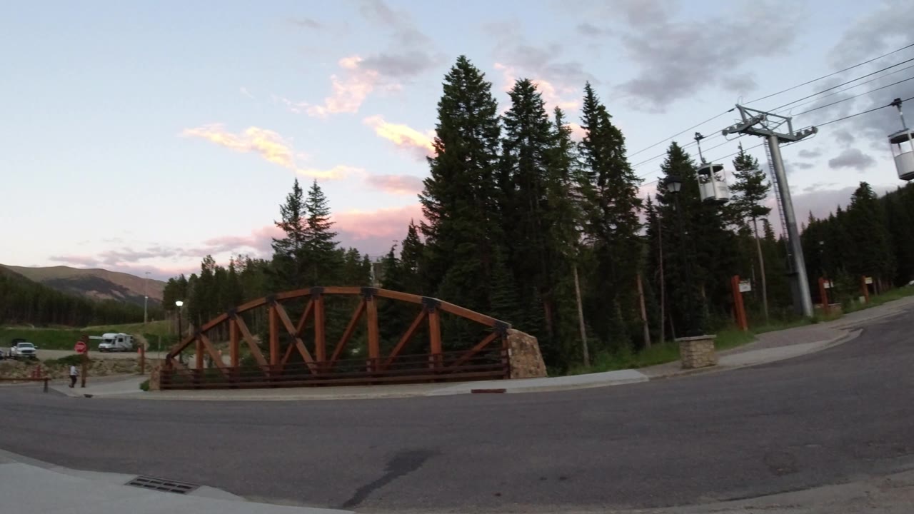 Winter Park village frame with the wooden bridge centered, a paved road in front, ski lift towers and cables overhead, parked cars in the distance, evergreen trees, and no close visible faces.