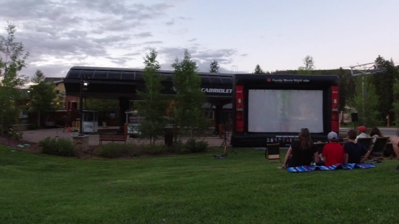 People sit on blankets on a grassy lawn in Winter Park, Colorado, facing an outdoor projection screen beside buildings and trees at dusk.