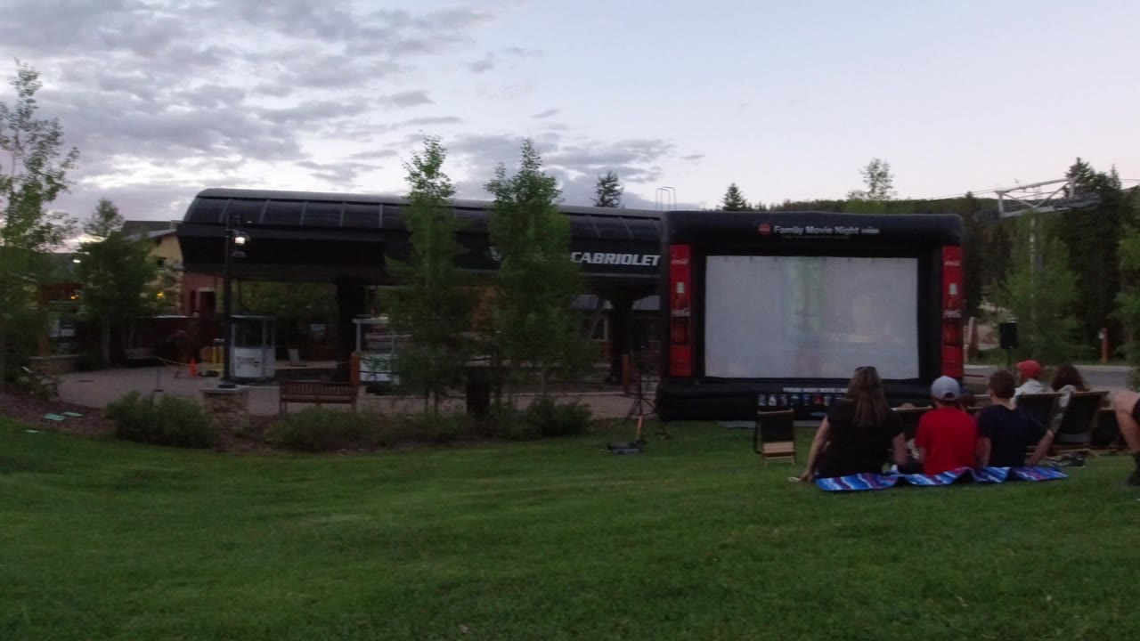 A seated group remains centered on the grass in Winter Park, Colorado, with the large outdoor screen, trees, and dark building facades behind them.