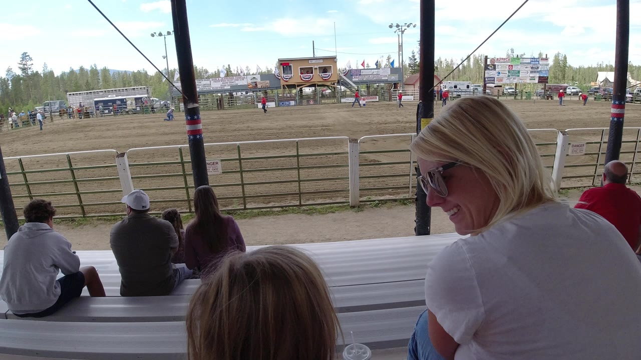 At 0:00 in Winter Park, Colorado, a woman and child sit in the foreground on bleachers while other spectators look toward a fenced dirt field and event buildings.