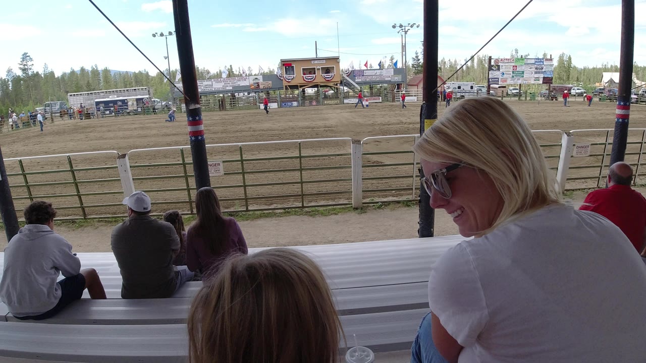 At 0:01 in Winter Park, Colorado, the foreground woman turns slightly on the bleachers as seated spectators continue watching the fenced field and distant event structures.