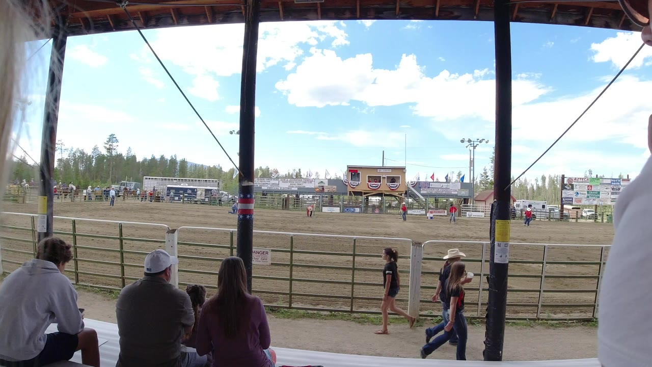 Spectator-level view in Winter Park, Colorado, with seated people under a shaded roof, a metal fence, a dirt arena, sponsor boards, and event buildings under bright sky.