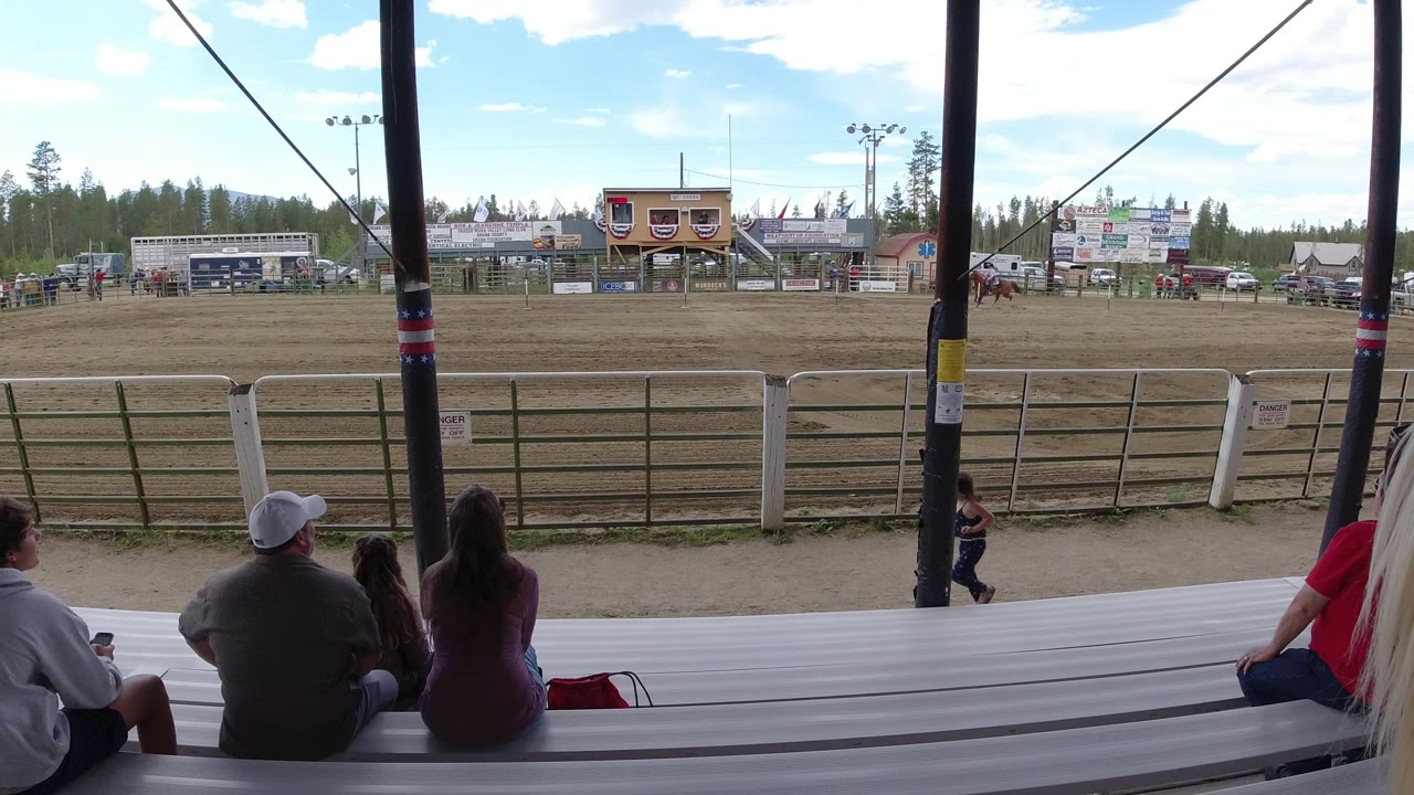 Wide covered-stand view in Winter Park, Colorado showing seated spectators on white bleachers, a fenced dirt horse track, riders in the distance, and arena buildings under bright daylight.