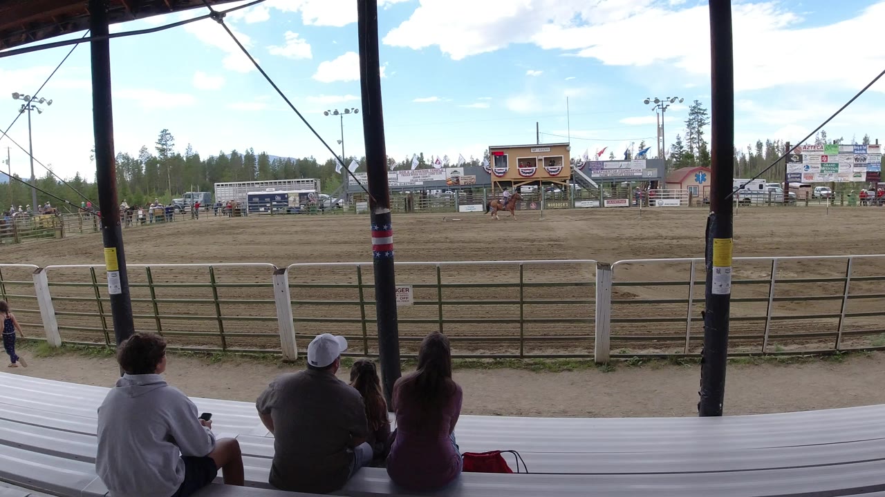 Spectators seen from behind sit under a covered stand in Winter Park, Colorado, facing a fenced dirt arena with event structures and trees in the background.