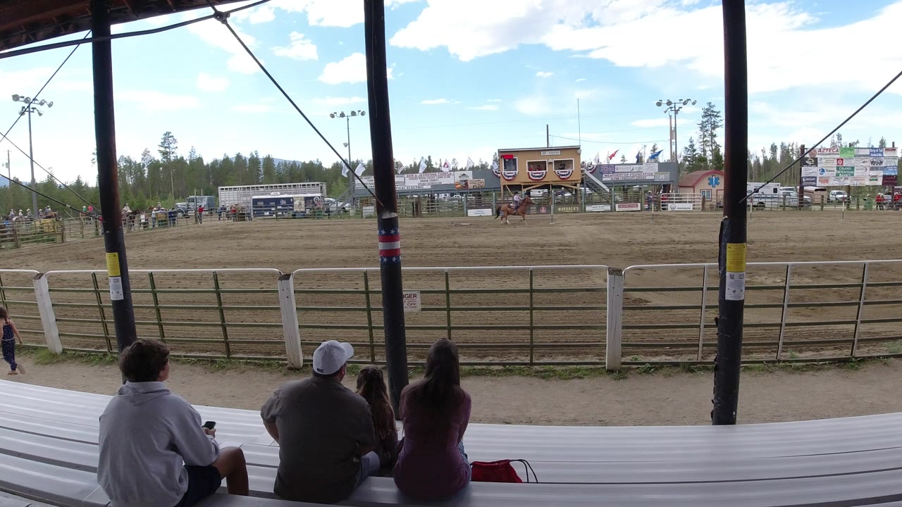 A small group of spectators remains seated in the foreground while the Winter Park, Colorado arena, fencing, signs, and tree line fill the distance.