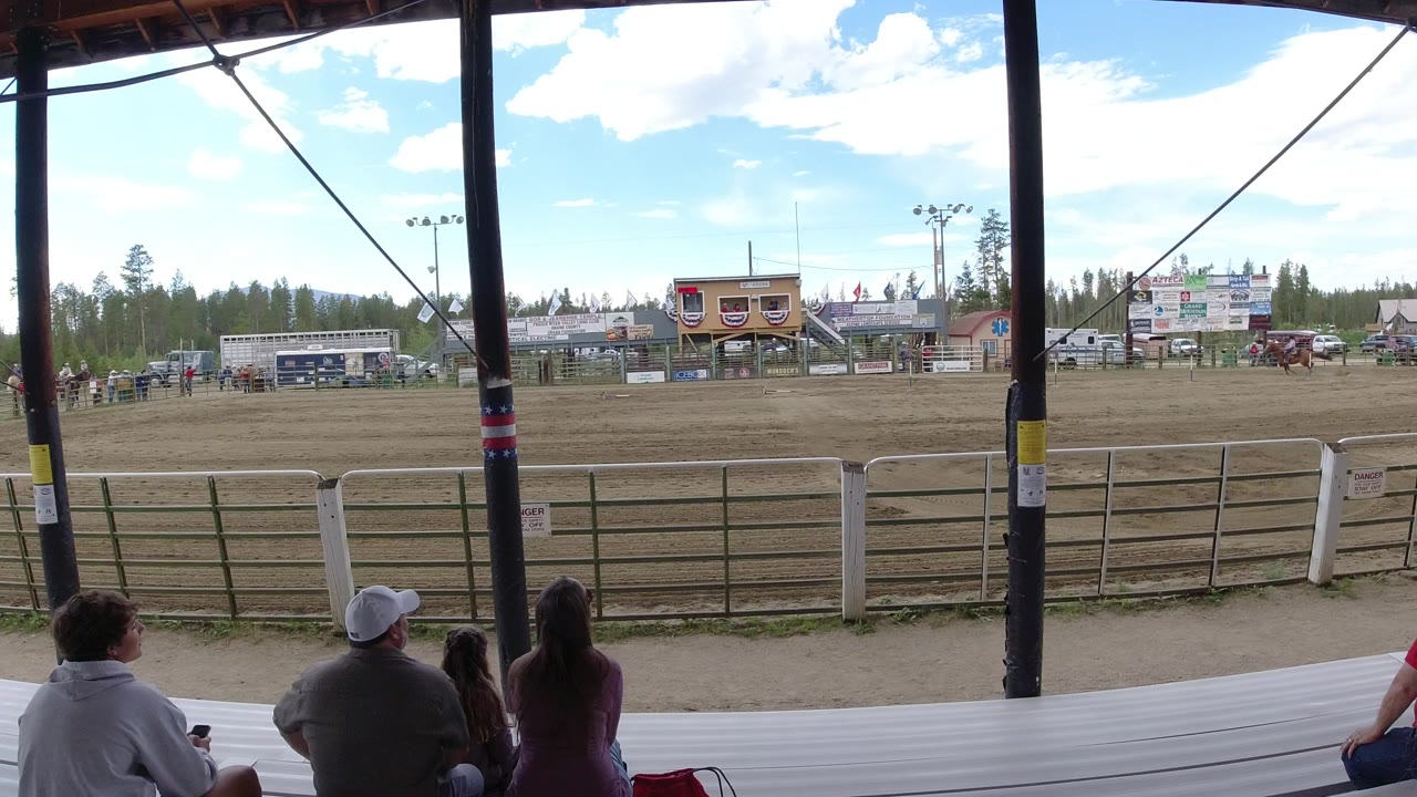 Spectators under a covered grandstand look toward a fenced dirt arena.