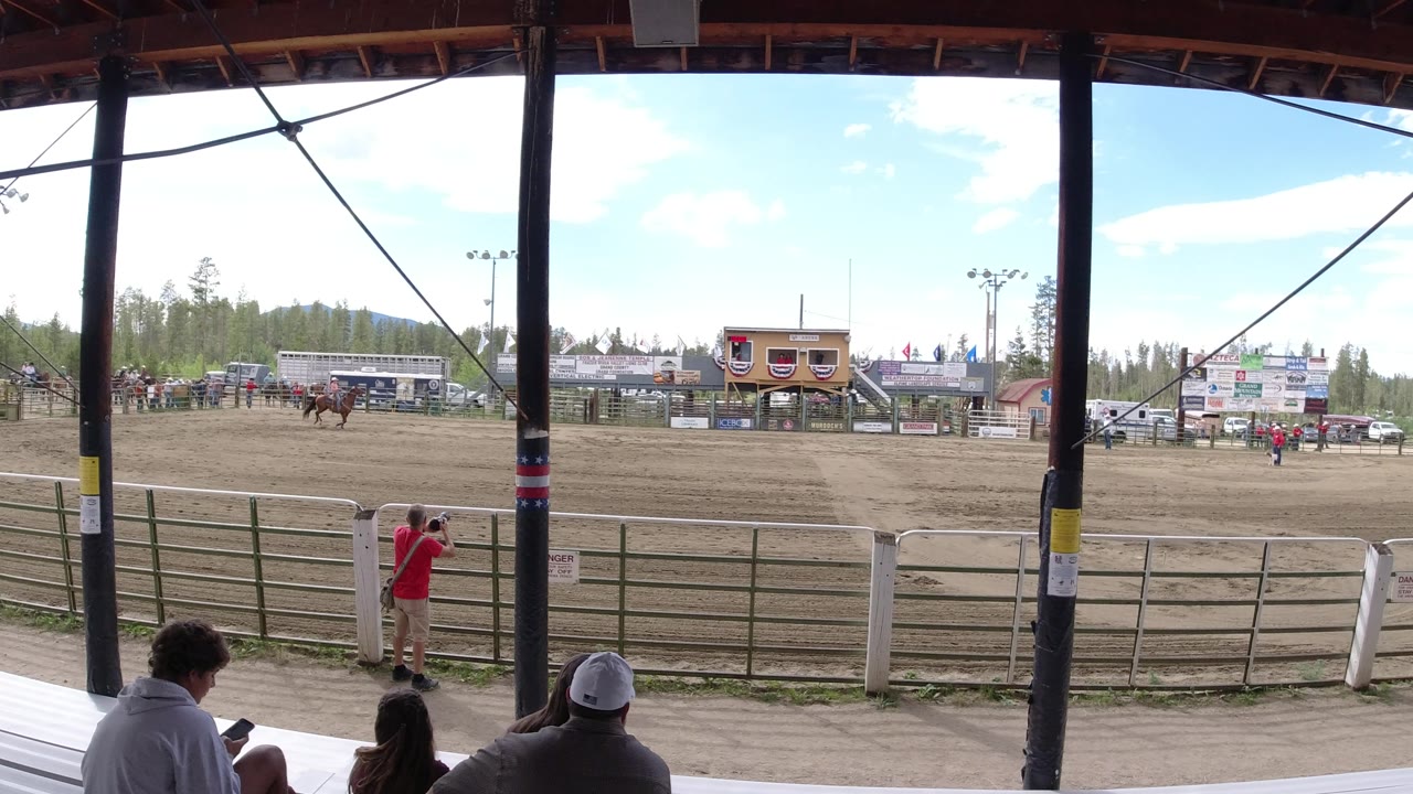 The Winter Park, Colorado arena remains framed by dark grandstand posts, with several spectators at the bottom edge, rail fencing across the foreground, and event structures and trees in the distance.
