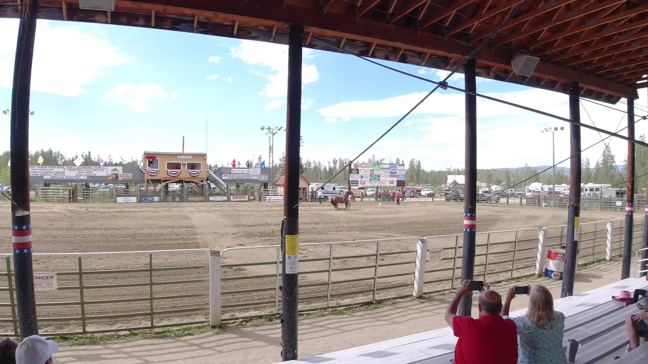 Wide opening view from covered bleachers in Winter Park, Colorado, with horses on the dirt arena, spectators in the foreground, fencing, signage, flags, trees, and hills beyond.
