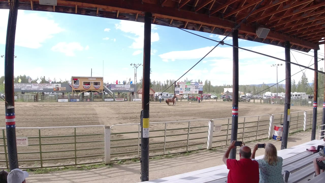 Horses continue across the Winter Park arena as two seated spectators in the foreground watch from metal bleachers beneath the covered structure.