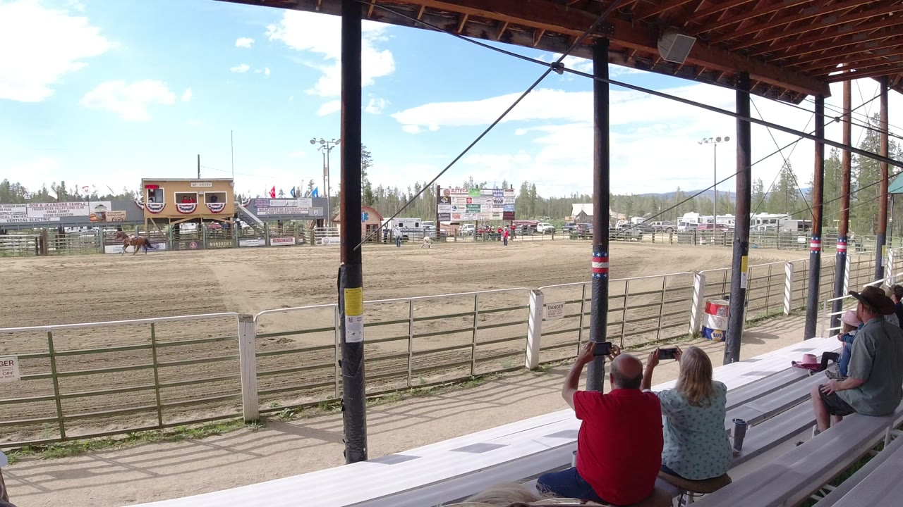 Spectators on white bleachers watch a dirt arena under a covered grandstand.