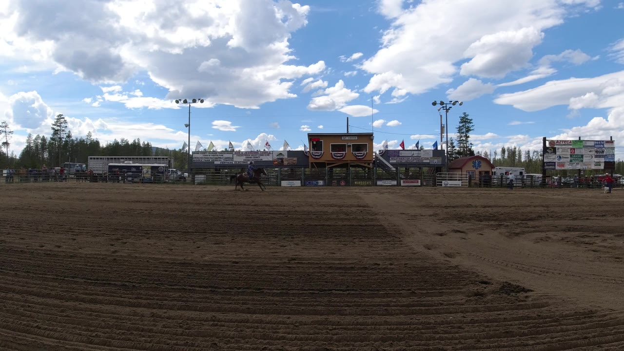Wide sandy arena with horses near bleachers and a scoreboard under cloudy sky.