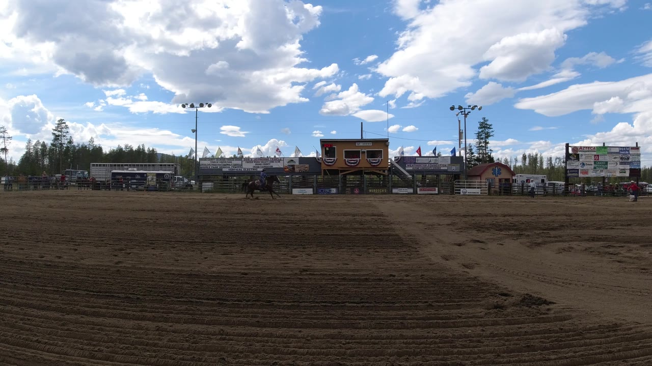In Winter Park, Colorado, a horse and rider appear near center frame on the sandy arena, backed by bleachers, advertising boards, flags, and a cloud-filled sky.