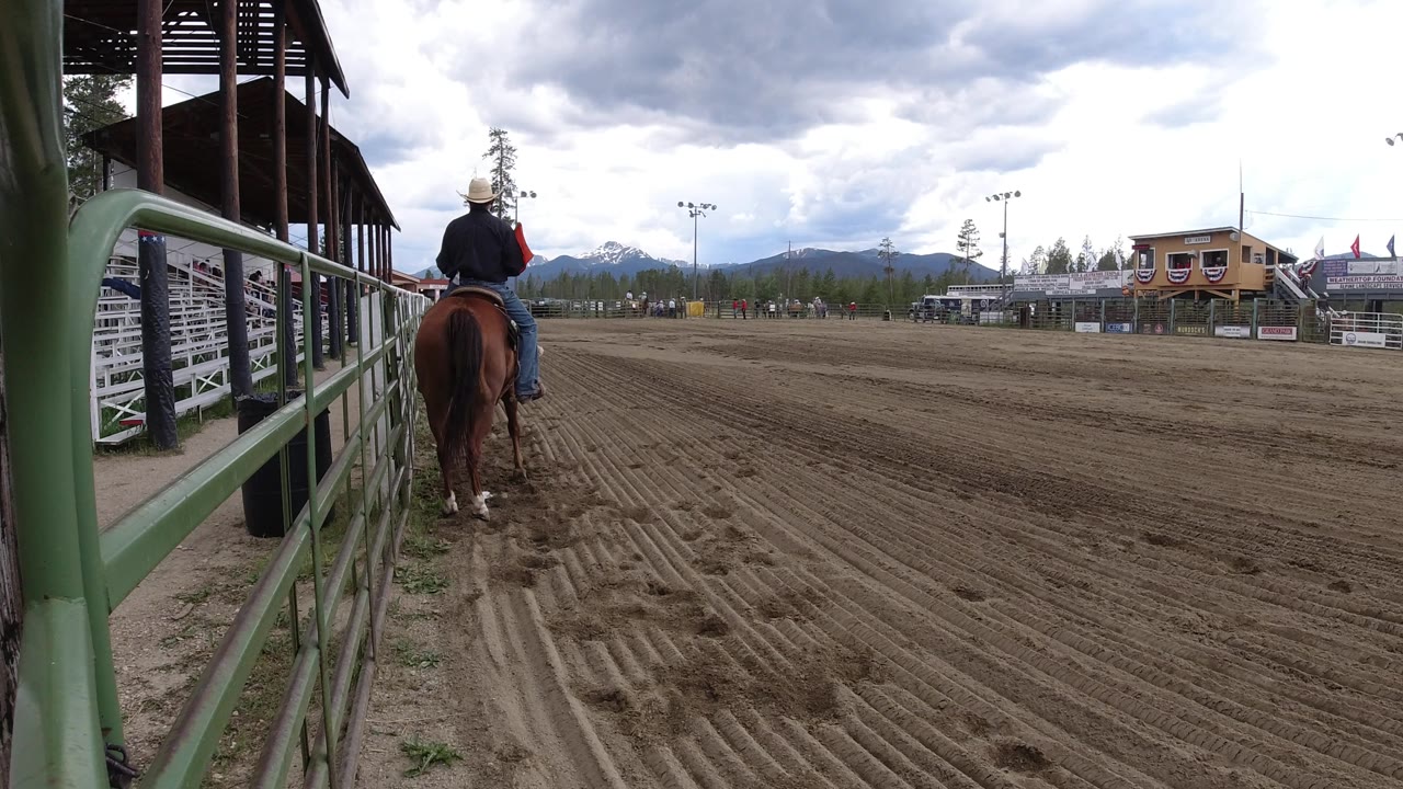 The mounted rider remains near the green rail in the Winter Park rodeo arena, with the dirt track opening wide toward bleachers and distant mountain ridges.