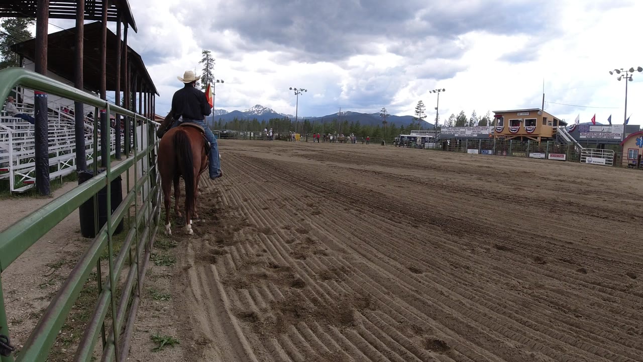 The horse and rider remain near the green fence in Winter Park, Colorado, while the open groomed arena, bleachers, small structures, and cloudy mountain horizon fill the frame.