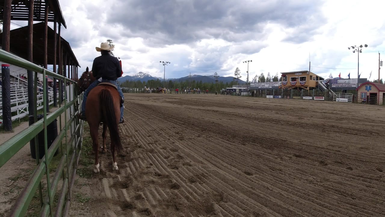 In Winter Park, Colorado, the rider remains mounted near the left rail while the broad raked dirt arena stretches toward small buildings and distant mountain slopes.